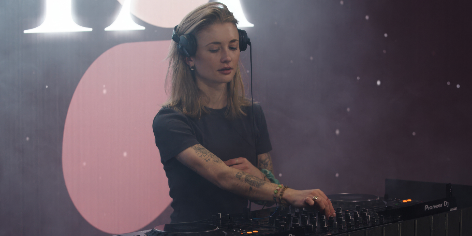 Caromelle a female DJ with tattoos adjusting mixer controls wearing headphones in a dim, smoky setting.