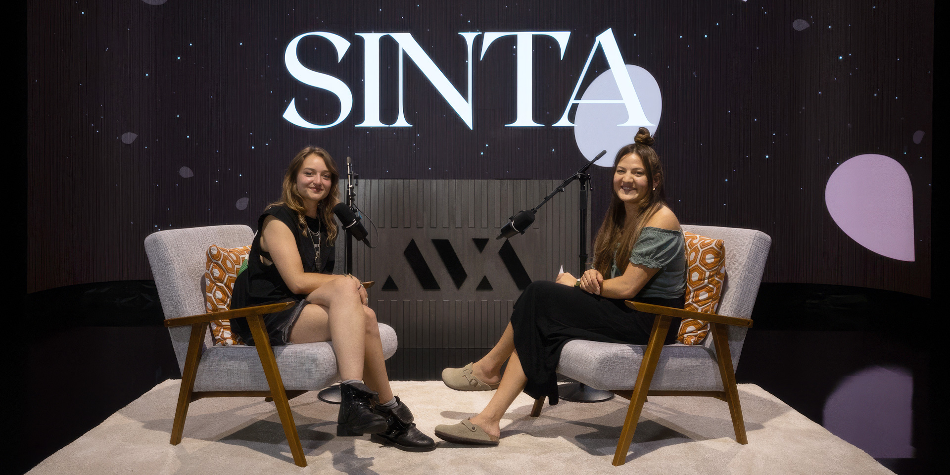 DJ Yamincy and Mia Soriano seated on armchairs in front of microphones with SINTA logo in background.