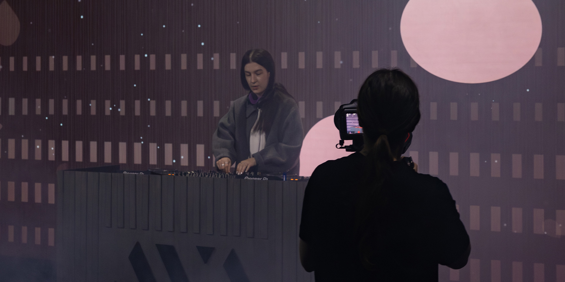 DJ performing on stage with a camera operator filming from behind, geometric shapes in background.