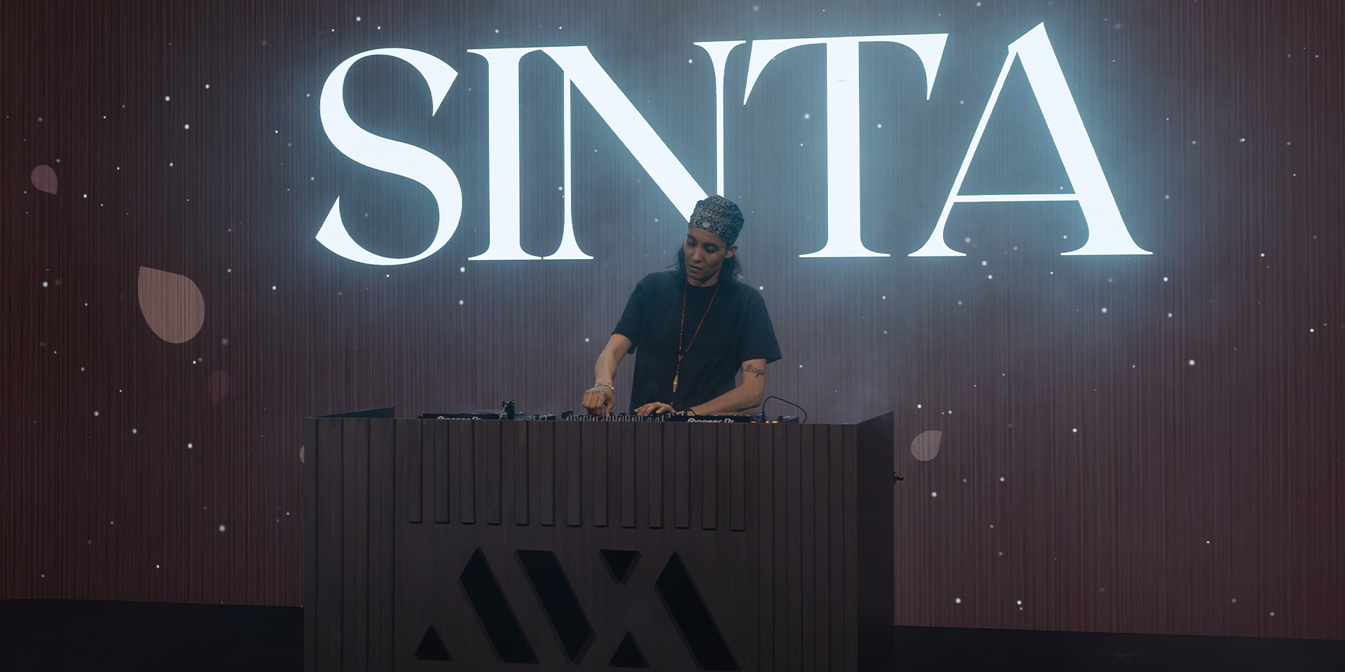 DJ wearing a patterned headband and black shirt mixing music on a console with SINTA lit behind.