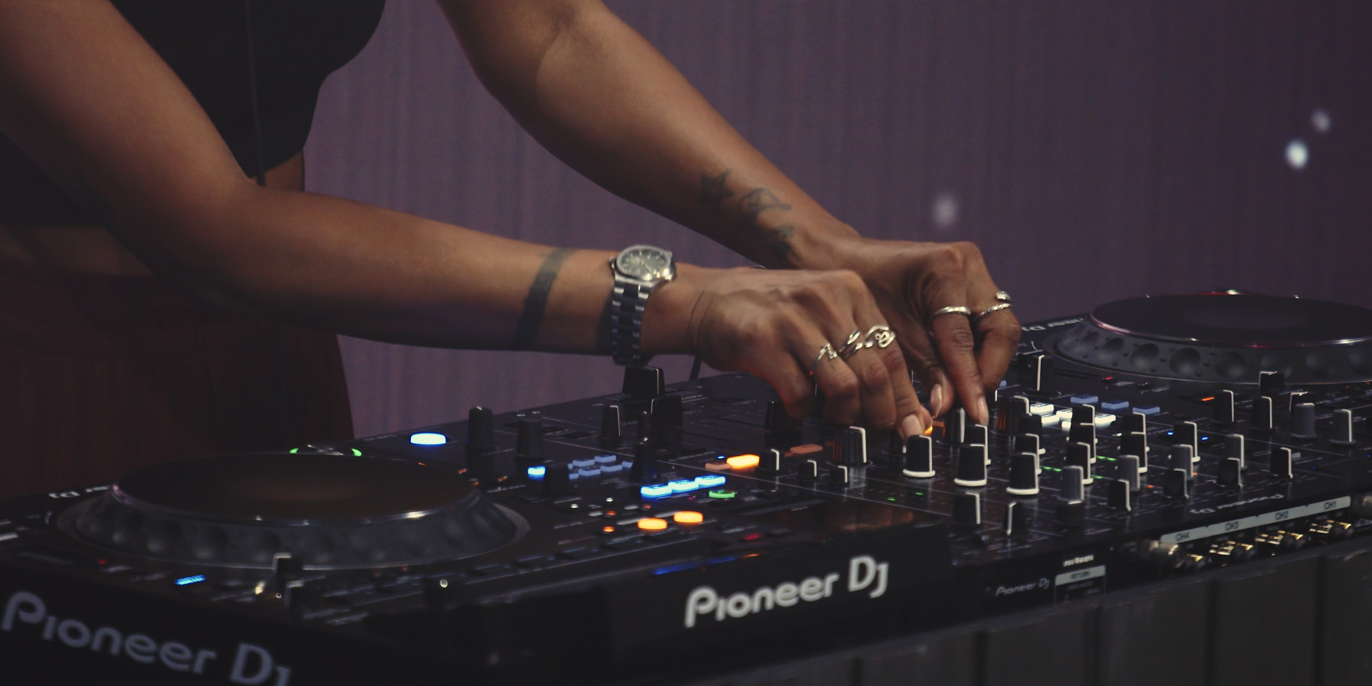 DJ adjusting knobs on a Pioneer DJ mixing console with tattoos and rings on hands visible.