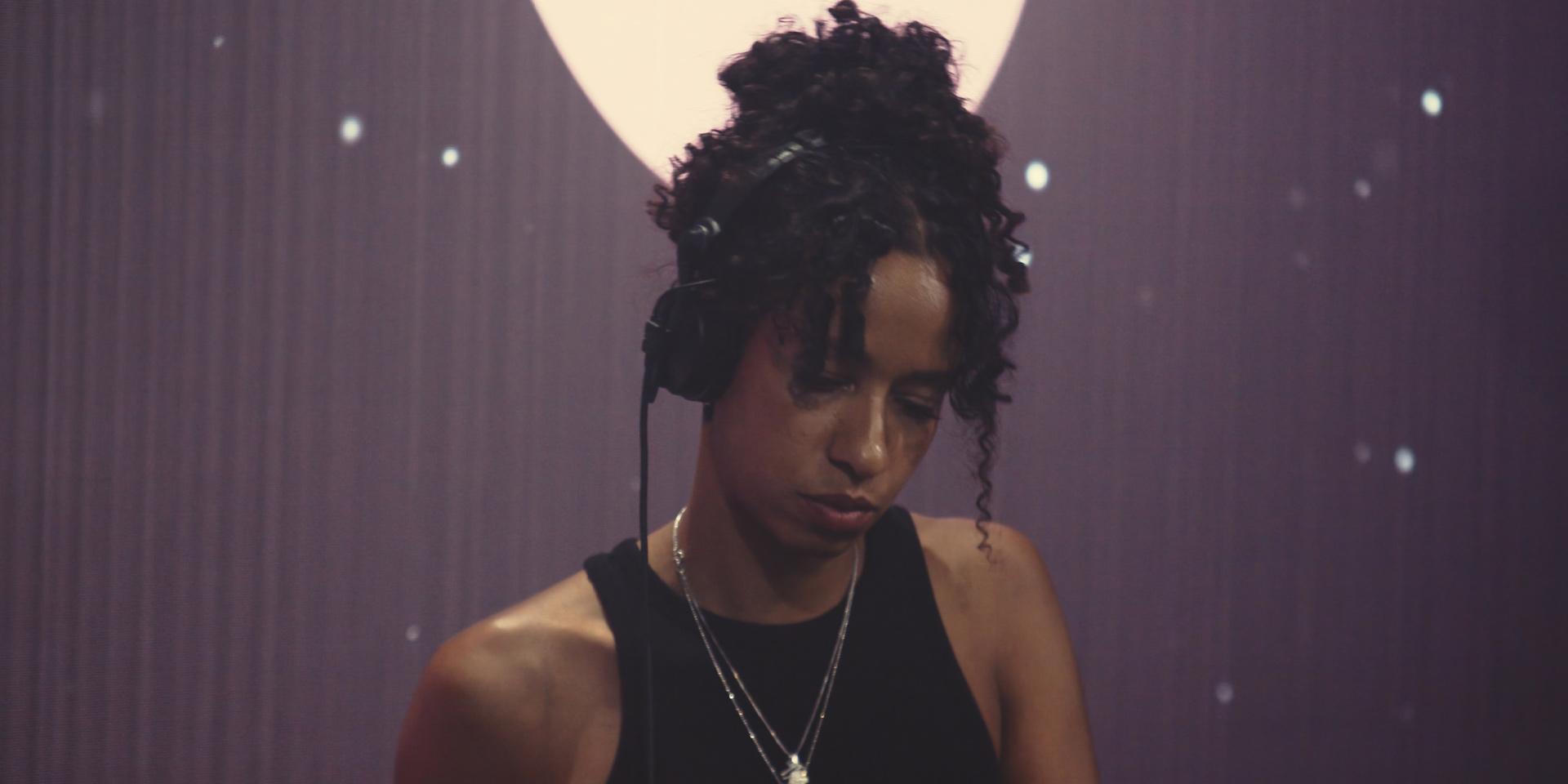 Woman DJ with curly hair wearing headphones and a black tank top, focused on her equipment.