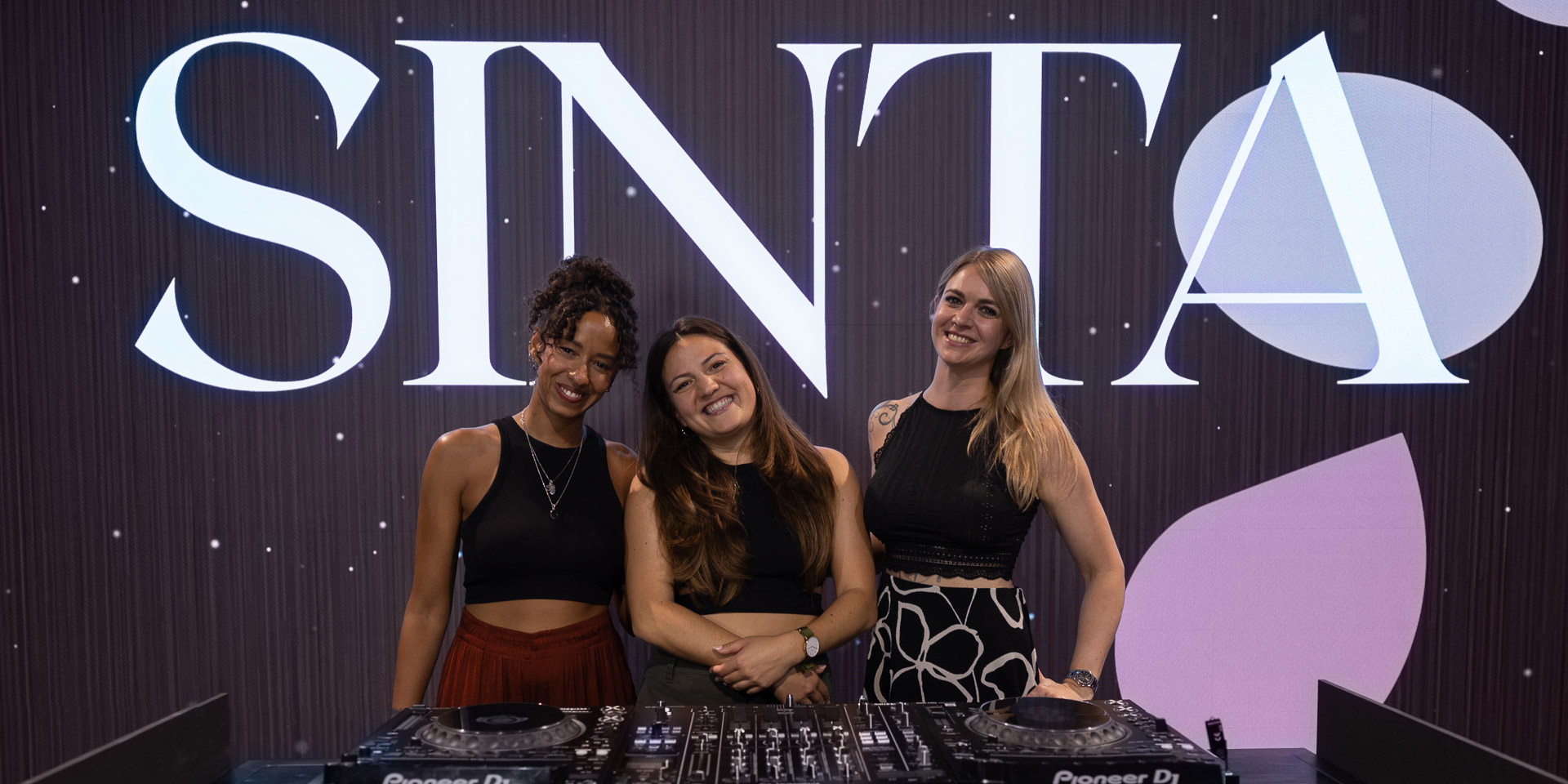 Three women smiling behind DJ mixer with large SINTA logo backdrop.