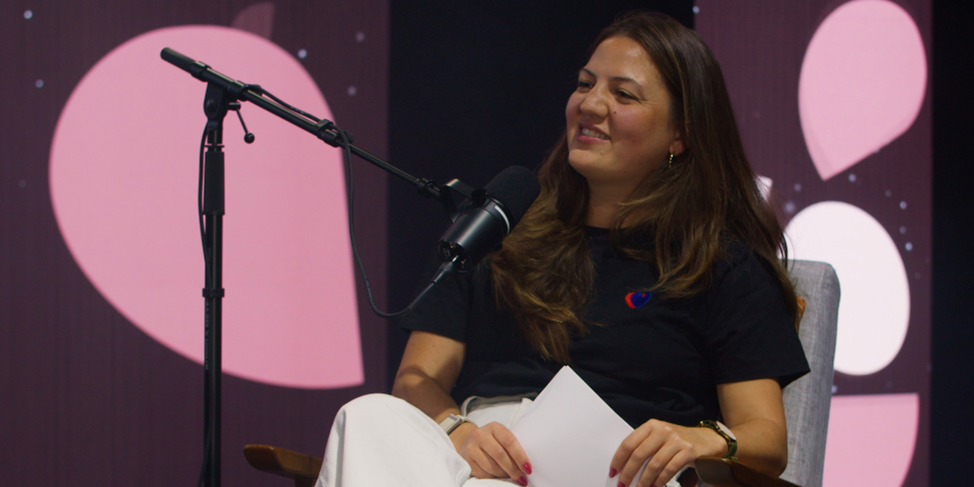 Woman smiling, sitting and holding paper, speaking into a microphone on a stand.