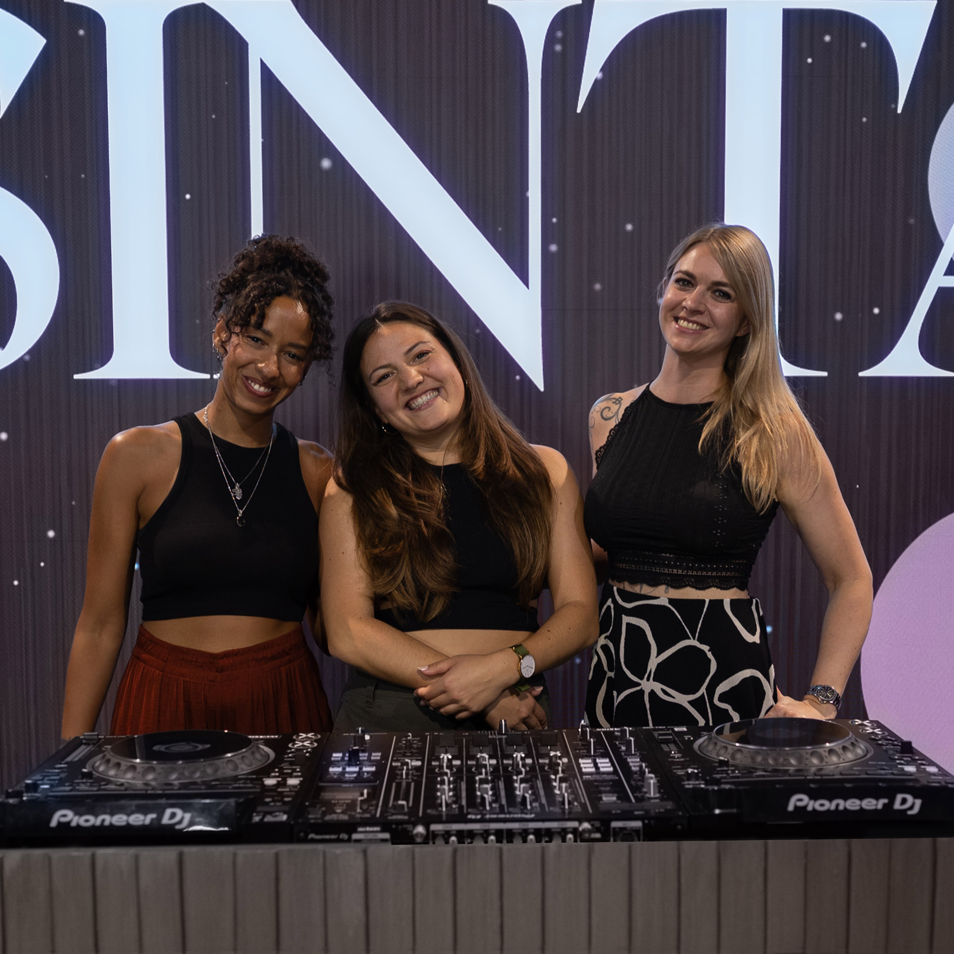 Three smiling women DJs standing behind Pioneer DJ equipment with a large lit sign behind them.