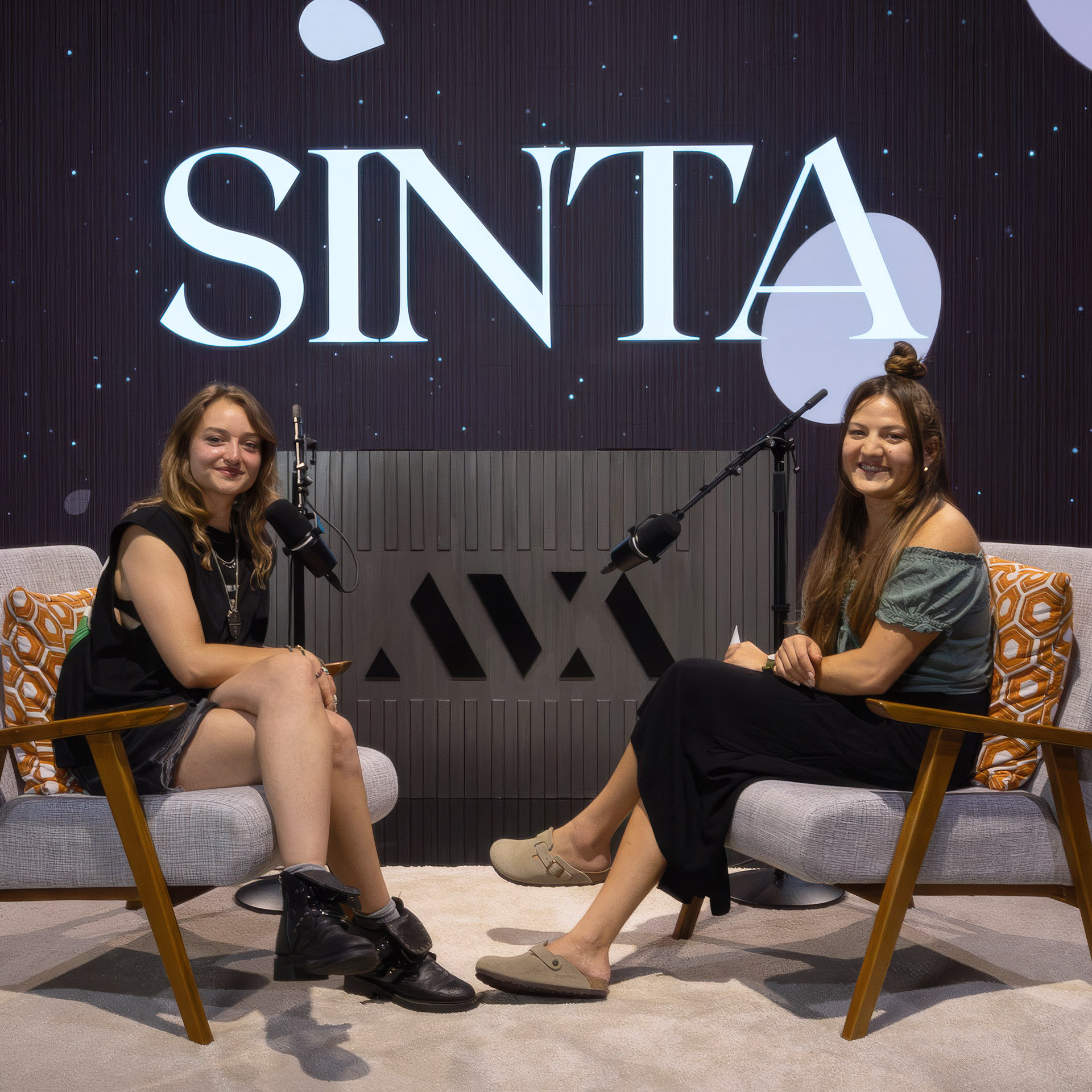 Two women seated in chairs facing each other with microphones and a 'SINTA' sign behind them.