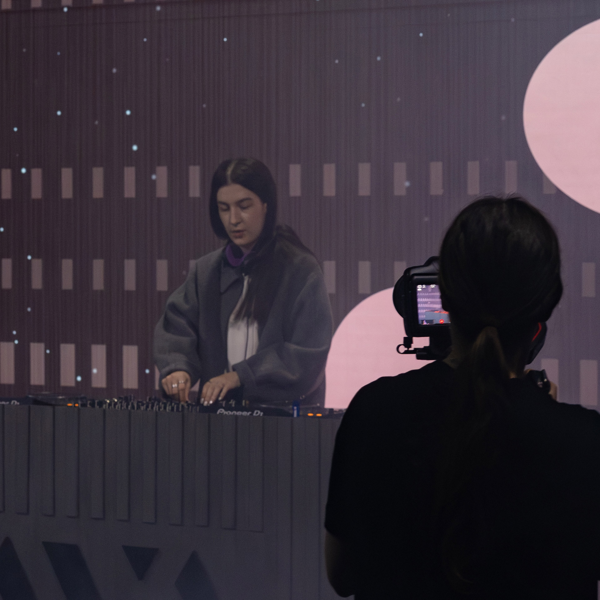 DJ wearing headphones performs on a Pioneer DJ set while being recorded by a camera operator.