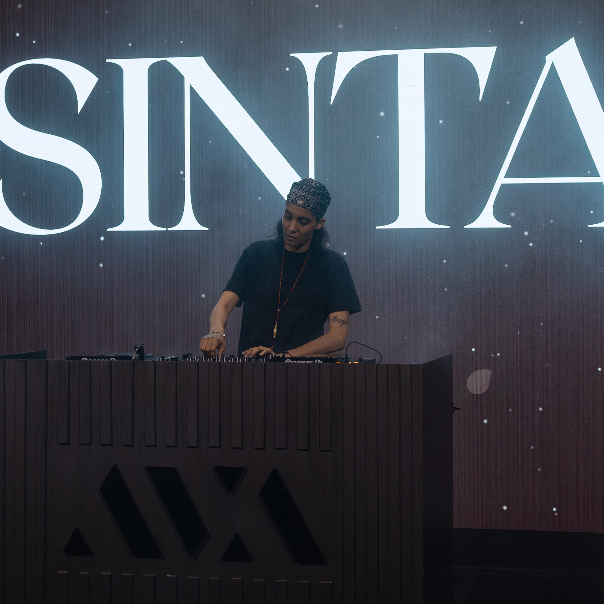 DJ wearing a bandana and black shirt mixing music on a console with large white SINTA letters behind.