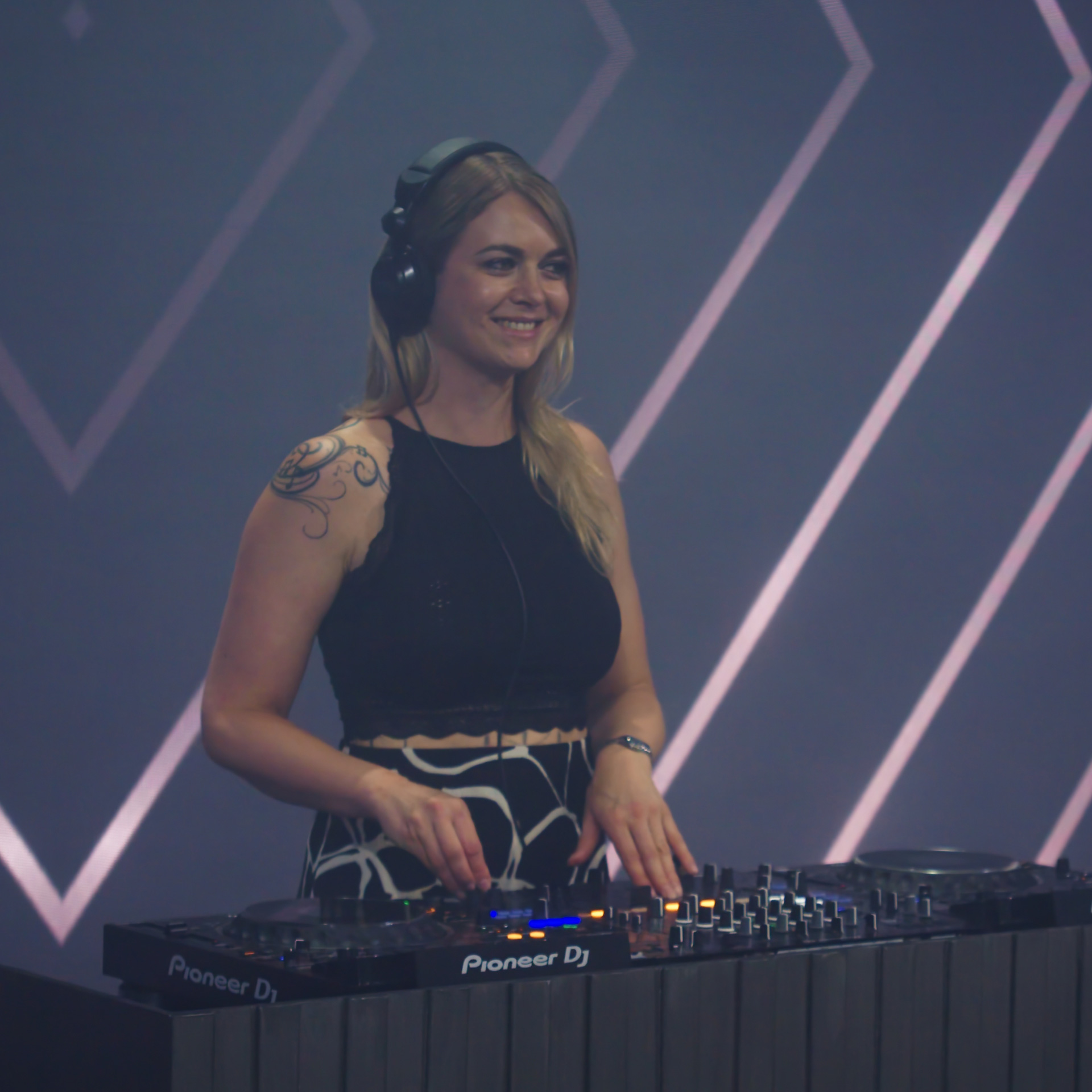 Smiling female DJ wearing headphones mixing music on a Pioneer DJ controller at an event.