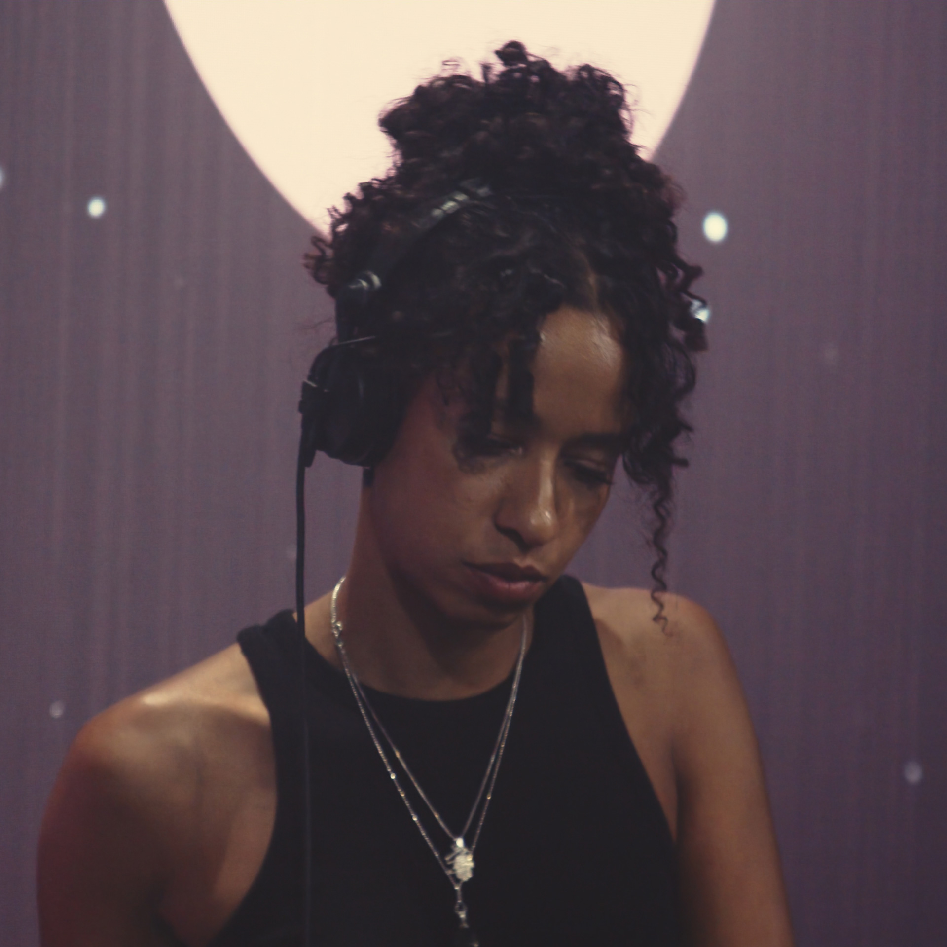 Woman with curly hair and headphones, wearing a black sleeveless top and layered necklaces, looking down.