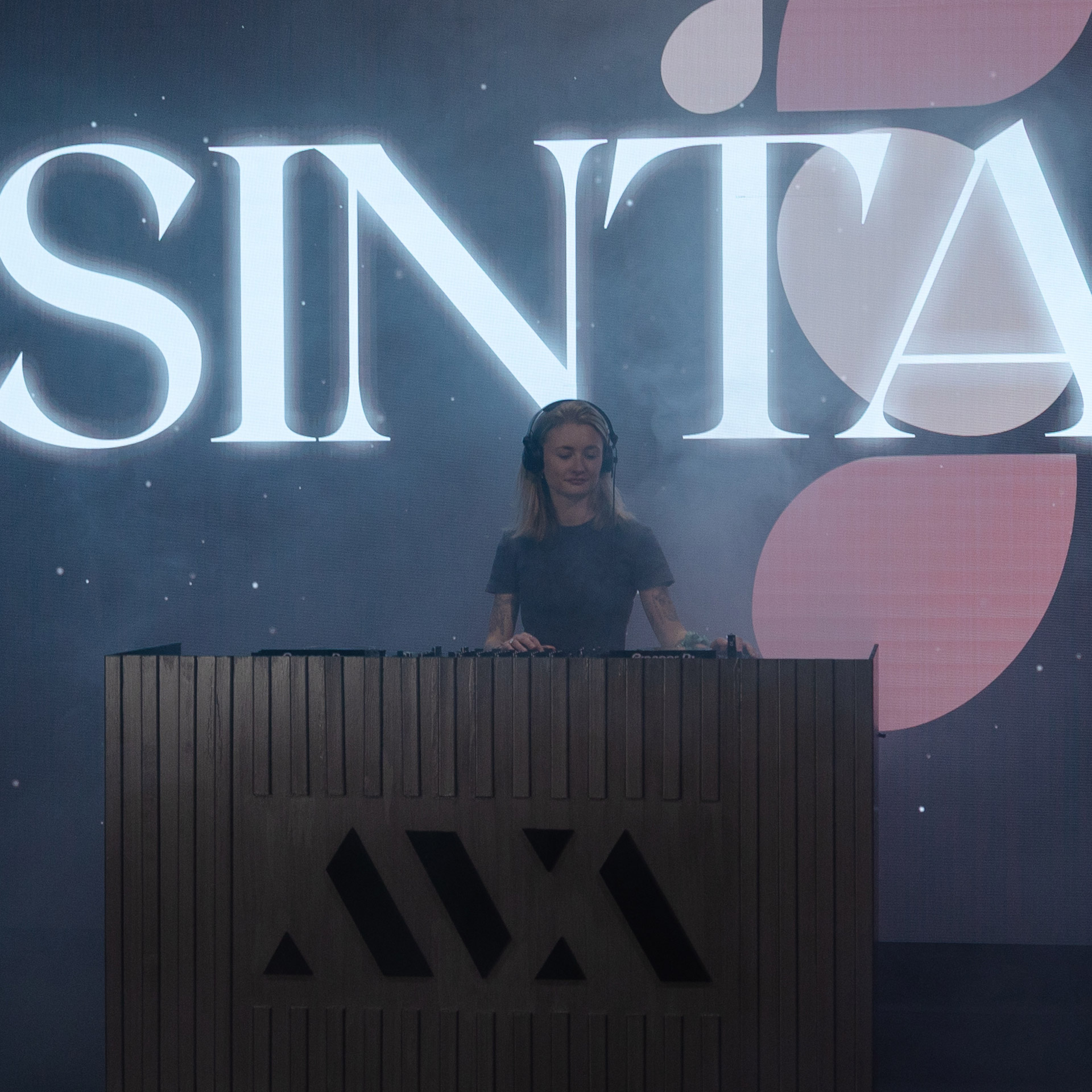 Female DJ wearing headphones performing behind a wooden booth with large 'SINTA' text behind her.
