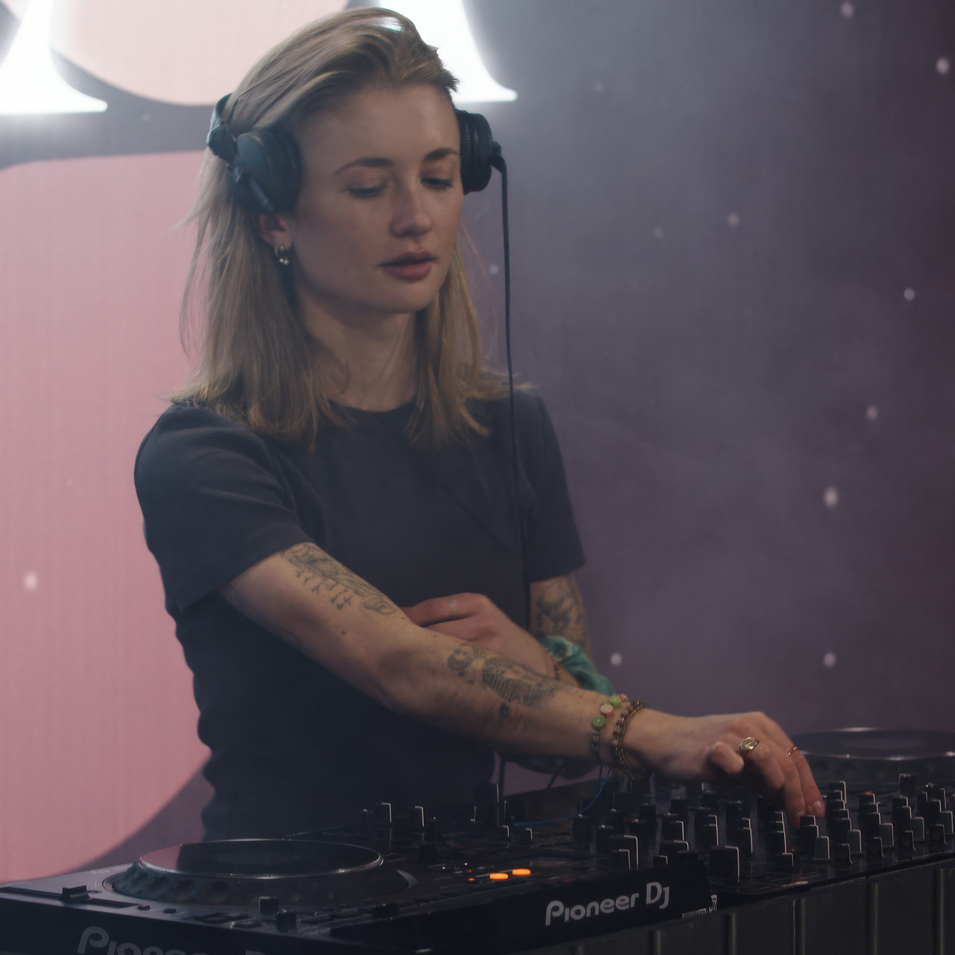 Female DJ with headphones using Pioneer DJ mixer, adjusting controls in a dimly lit space.